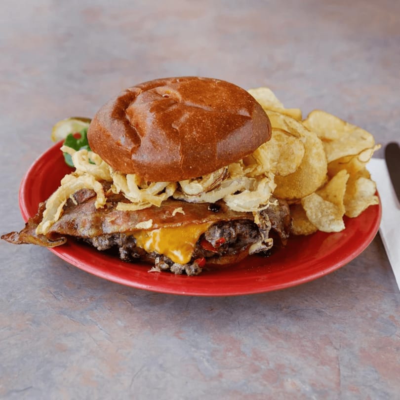 A loaded burger with bacon, cheese, and crispy onions, served with potato chips and pickles.