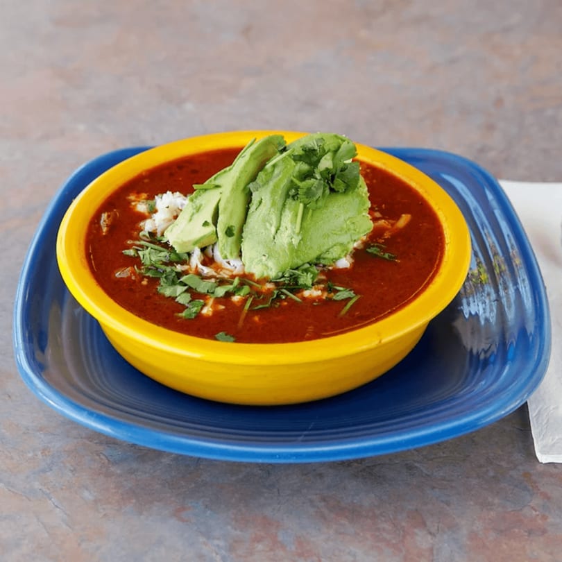 Red soup in a yellow bowl, topped with avocado slices, cilantro, and white cheese, on a blue plate.