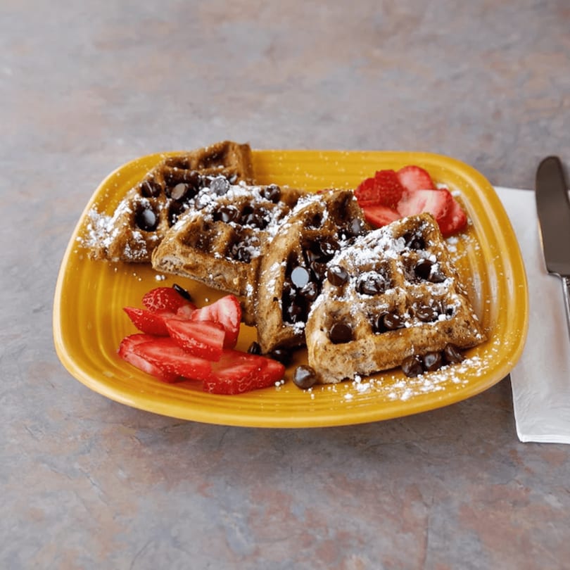 Chocolate chip waffles with powdered sugar and sliced strawberries on a yellow plate.
