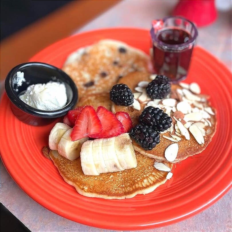 Pancakes with strawberries, bananas, blackberries, almonds, whipped cream, and syrup on a red plate.